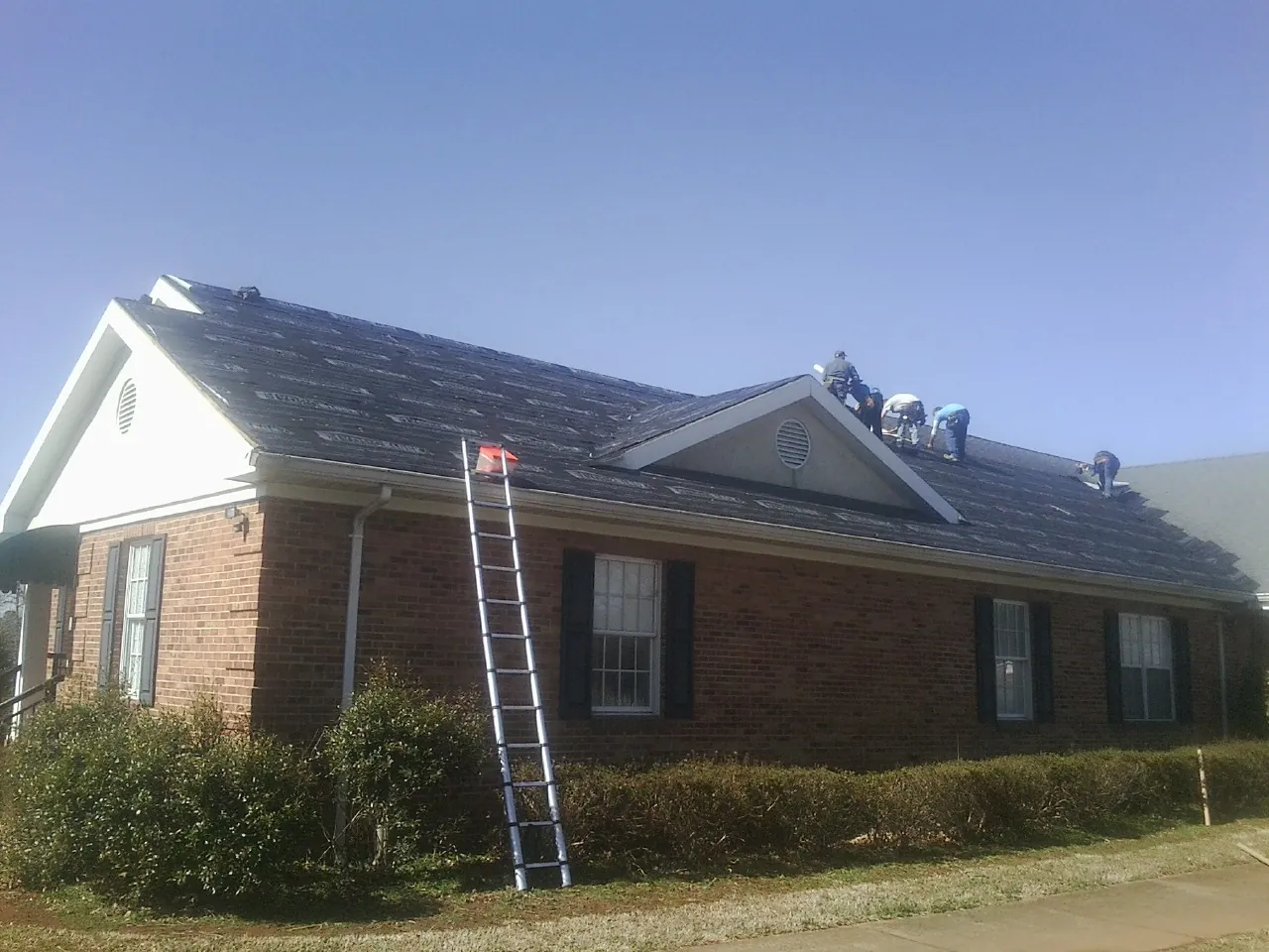 Crew tearing shingles off a brick ranch roof with a ladder set — Emergency Roof Repair work by our crew in Gratiot