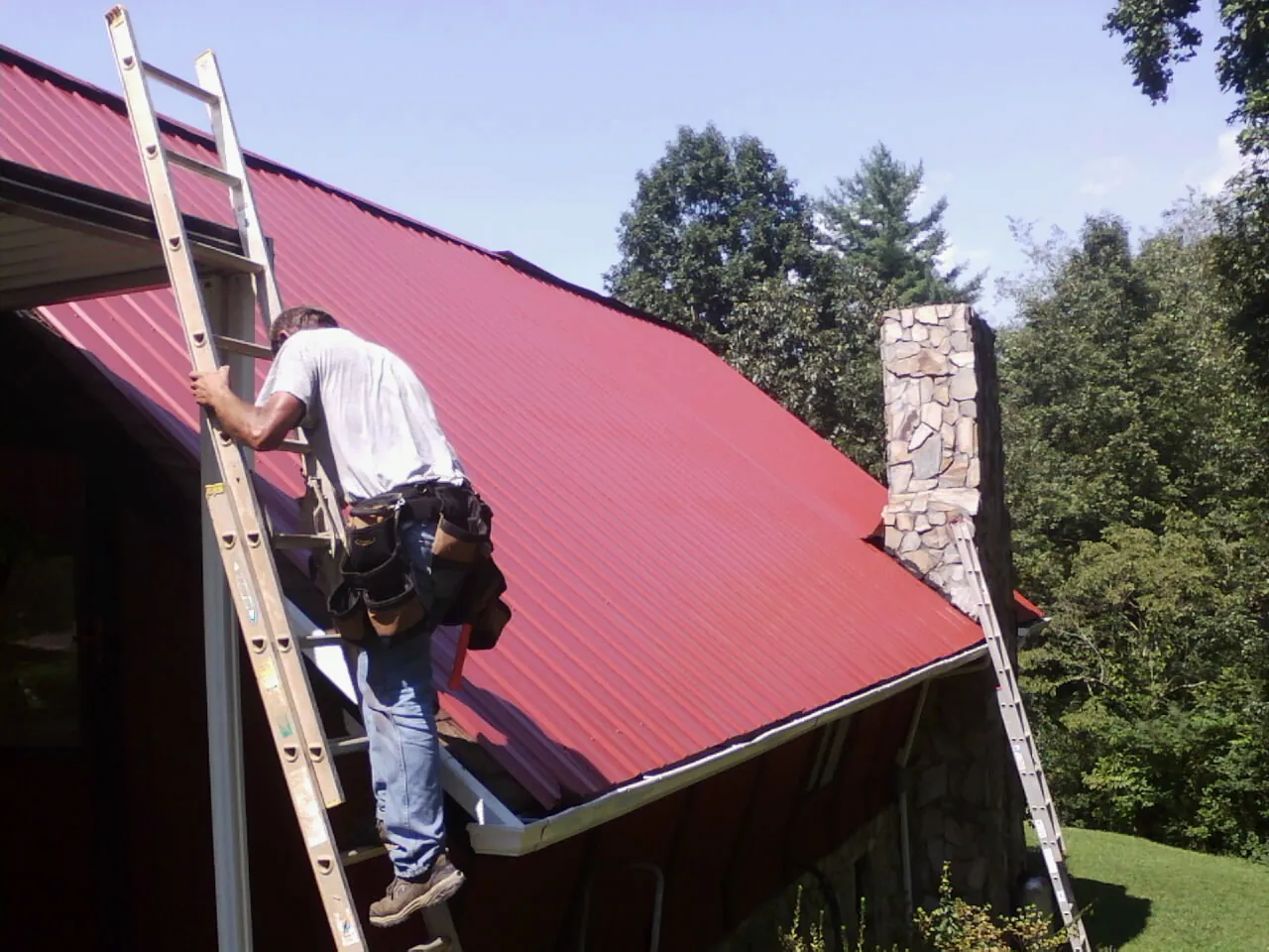 Roofer on a ladder beside a red metal roof and stone chimney — Soffit & Fascia Repair by Roofing Brothers in Gratiot